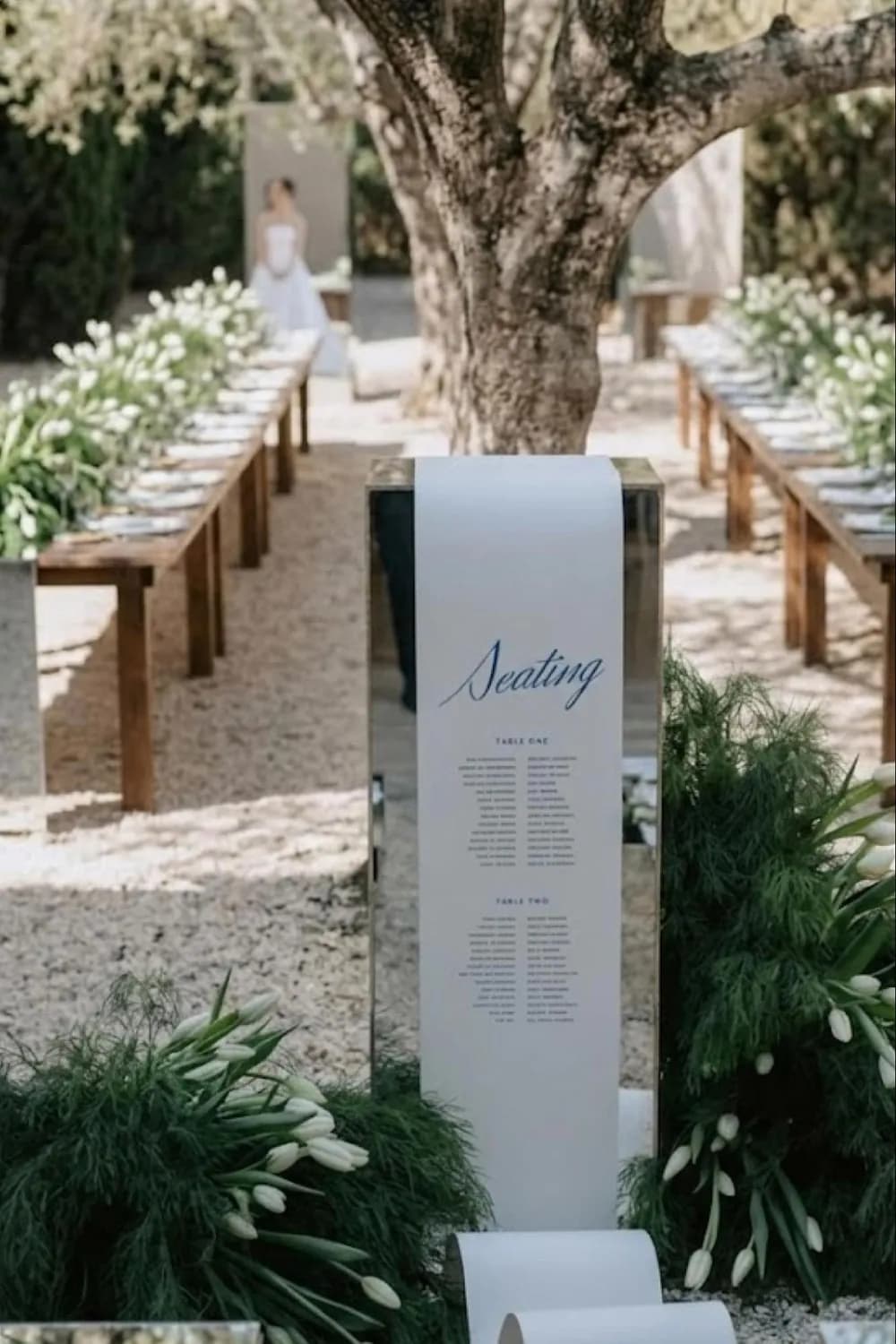 A white 'Seating' calligraphy board lining a tree-shaded outdoor aisle, its base planted with greenery