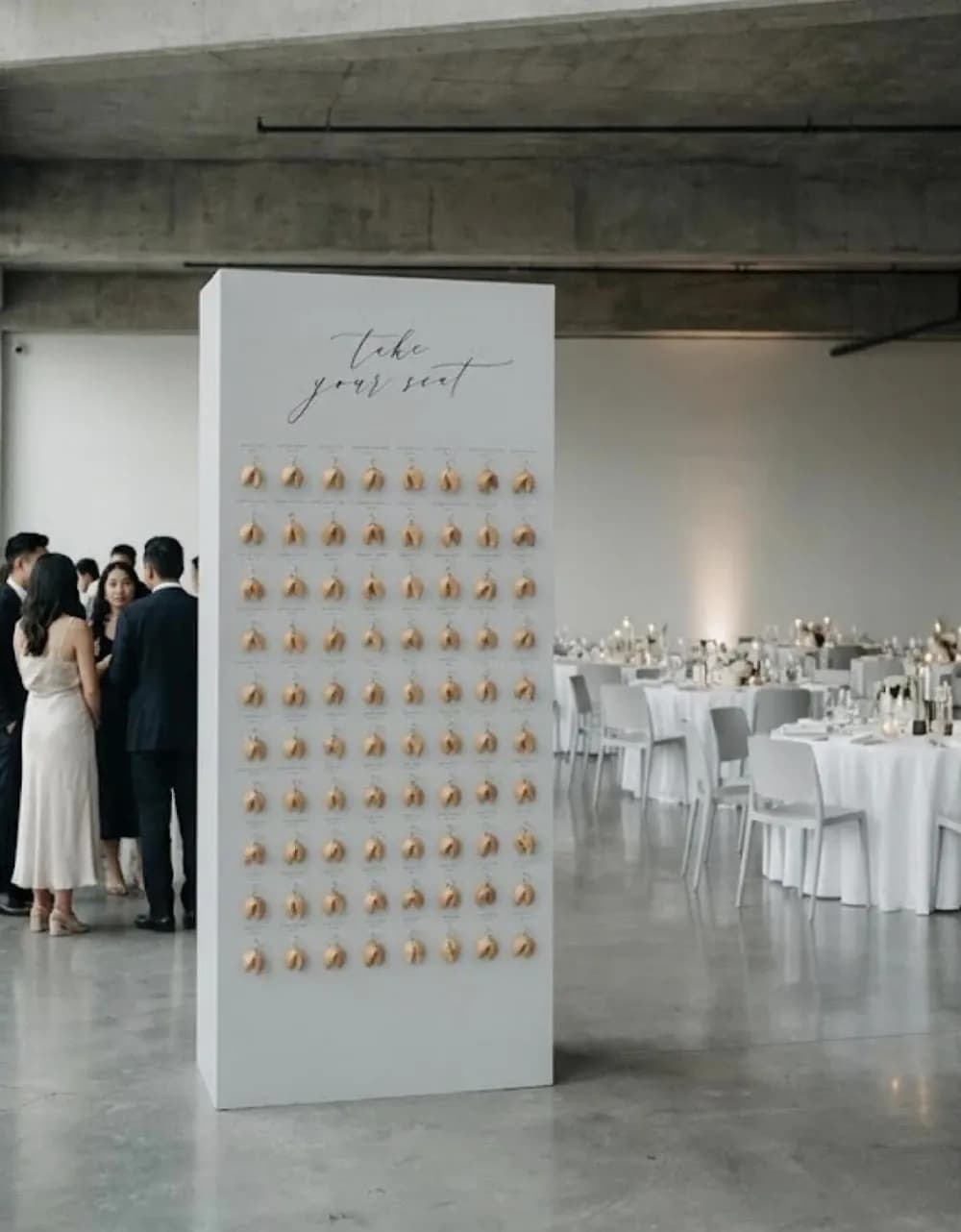 A tall white fortune-cookie seating wall in a concrete loft venue, a grid of cookies under 'take your seat' calligraphy