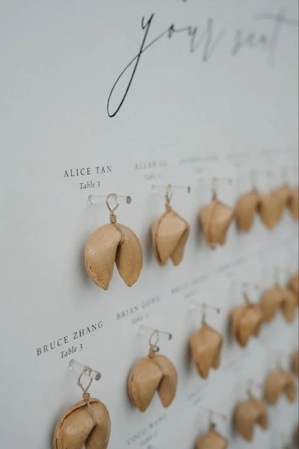 Fortune-cookie wedding seating chart: real cookies hang from clear pegs on a crisp white board, each beside a guest name and table number