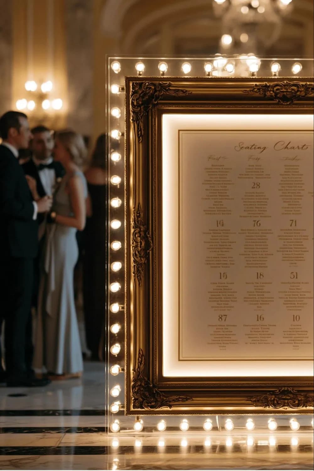 Hollywood-glam wedding seating chart: an ornate gilt frame ringed in glowing marquee bulbs around a backlit chart with names in script, in a chandelier-lit ballroom