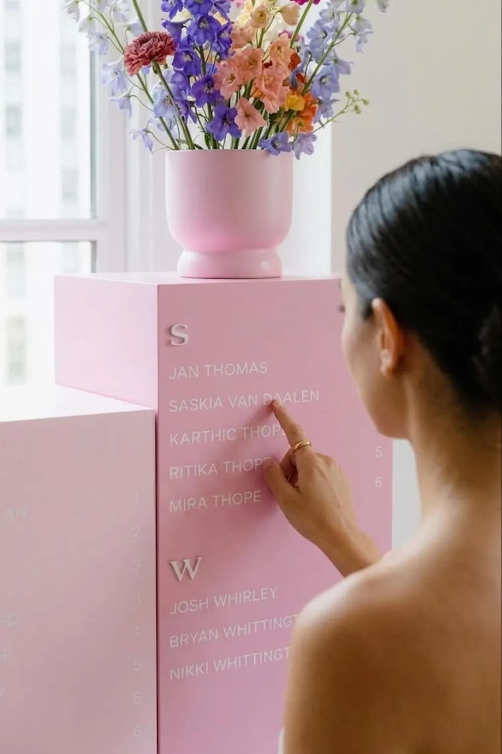 A blush-pink plinth column seating chart with names grouped by surname initial, styled with a flower arrangement on top