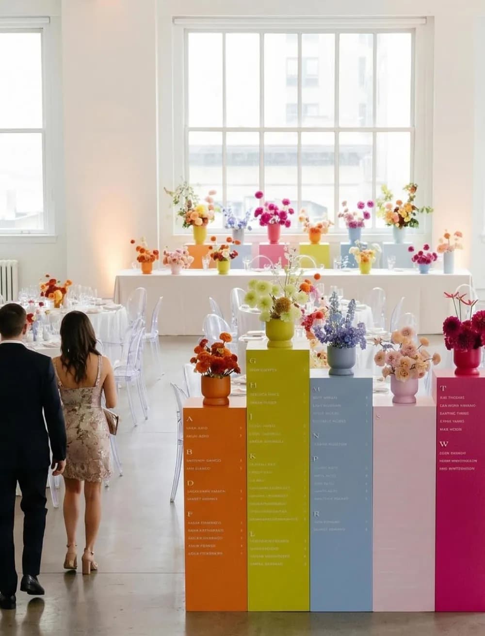 A row of saturated plinth columns in orange, lime, blue, pink and magenta forming a gradient seating chart, topped with bright florals in a loft