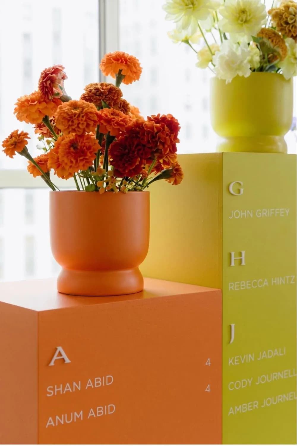 Minimalist seating chart on orange and chartreuse plinth columns with alphabetical names, topped with marigolds in coordinating vases